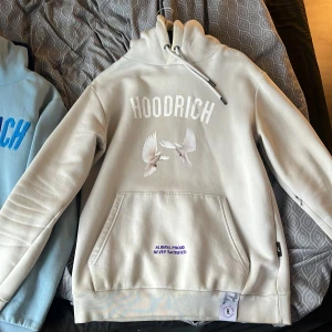 Vit hoodie från Hoodrich med broderade detaljer - Säljer en vit hoodie från Hoodrich med broderad text och motiv av duvor på både fram- och baksidan. Hoodien har känguruficka, dragsko i huvan och tryck i lila och vitt. Snygga detaljer som texten 'From Nothing To Something' och 'Always Proud Never Satisfied'.