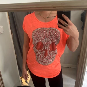 Neonorange t-shirt med dödskalletryck - Cool neonorange t-shirt med stort glittrigt dödskalletryck framtill. Rund halsringning och korta ärmar. Perfekt statement-plagg för dig som vill sticka ut.