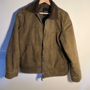 Olivgrön jacka med dragkedja från Monki - Säljer en olivgrön jacka från Monki med två dragkedjor framtill och två stora fickor. Jackan har klassisk krage och lång ärm, perfekt för lager-på-lager. Snygg och enkel design som passar till mycket.