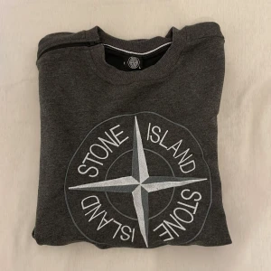 Grå sweatshirt från Stone Island - Säljer en grå sweatshirt från Stone Island med stort broderat logomärke på bröstet och klassisk patch på ärmen. Tröjan har rund halsringning och ribbade muddar vid ärmslut och nederkant. Perfekt för en stilren och avslappnad look.