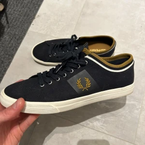 Mörkblå sneakers från Fred Perry - Säljer ett par mörkblå Fred Perry sneakers med vit sula och gulddetaljer vid kragen. Skorna har snörning och är tillverkade i textil med Fred Perrys ikoniska lagerkrans på sidan. Perfekta för en stilren och sportig look. Nypris 1799:-
