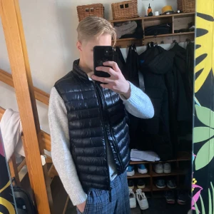 Svart dunväst från Peak Performance - Snygg svart dunväst från Peak Performance med glansig finish och ståkrage. Västen har en klassisk passform och är perfekt att bära över en tröja för extra värme. Diskret logotyp på bröstet och dragkedja framtill.