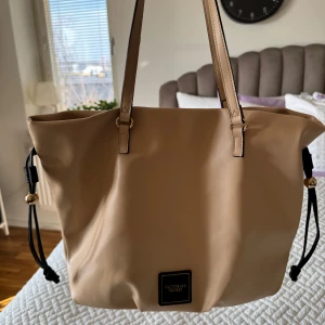 Victoria’s Secret väska - Säljer min mors Victoria’s Secret väska.  Inköpt för ca 2 år sedan som present. Aldrig använd. Inköpspris 700kr. Banden på sidan spänns åt och på så sätt stänger man till väskan.