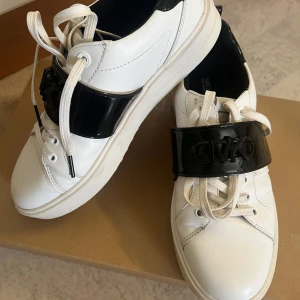 Vita sneakers från Michael Kors med svart detalj - Säljer ett par vita sneakers från Michael Kors med bred svart lackdetalj och MK-logga över vristen. Skorna har vita snören, rund tå och platt sula. Perfekta för dig som gillar stilrena men ändå iögonfallande skor.