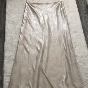 Champagnefärgad långkjol från Gina Tricot - Säljer en glansig, champagnefärgad långkjol från Gina Tricot. Kjolen har diskret struktur i tyget och stängs med en dragkedja i sidan. Perfekt för dig som gillar en elegant och stilren look.