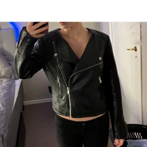 Svart skinnjacka med dragkedjor - Säljer en svart skinnjacka med silvriga dragkedjor och klassisk bikerstil. Jackan har snedställd dragkedja framtill, fickor med dragkedja och detaljerade sömmar på axlarna. Perfekt för en cool och stilren look.