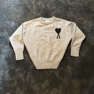 Grå stickad tröja från Ami Paris - Säljer en stilren grå stickad tröja från Ami Paris med svart hjärta och bokstaven A broderat på bröstet. Tröjan har rund halsringning och långa ärmar med ribbade muddar och nederkant.