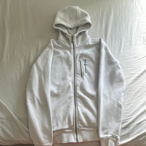 Vit hoodie från Sail Racing - Vit hoodie från Sail Racing med dragkedja framtill och en extra dragkedjeficka på bröstet. Broderad logga på bröstet och justerbar huva med snörning. Perfekt för en sportig och stilren look. Andväd 10-15 gnr.