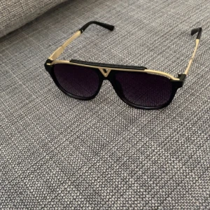 Svarta och guldiga LV solglasögon med  - Snygga solglasögon med svart båge och guldiga detaljer. Glasen är lila tonade och modellen har en markerad överdel. Perfekt accessoar för att lyfta din stil. Prisk kan diskuteras 