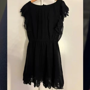 Svart volangklänning från KappAhl - Supersöt svart klänning från KappAhl med volangdetaljer på ärmarna och spetskant nertill. Klänningen har en markerad midja och är tillverkad i ett lätt, flowy material. Perfekt för dig som gillar en klassisk look med fina detaljer.