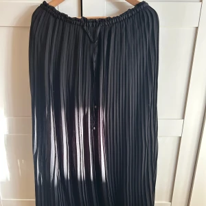 Svart plisserad långkjol - Säljer en svart plisserad långkjol med resår i midjan. Kjolen har ett snyggt fall och är tillverkad i ett lätt, glansigt material som ger en elegant känsla. Perfekt att styla med en enkel topp eller stickad tröja.