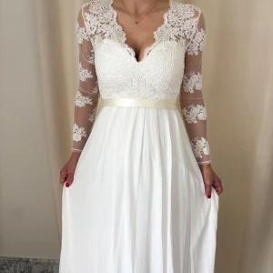  Bröllop långklänning med spets och rosett - Romantisk vit långklänning med v-ringad spetsliv, transparenta spetsärmar och öppen rygg. Klänningen har ett satinband i midjan som knyts till rosett bak och en mjukt fallande kjol. Perfekt för speciella tillfällen.