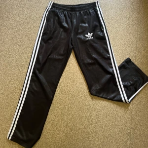 Adidas byxor - Glansiga svarta Adidas byxor med vita ränder längs sidorna och broderad logga framtill. Ena dragkedjan är sönder 
