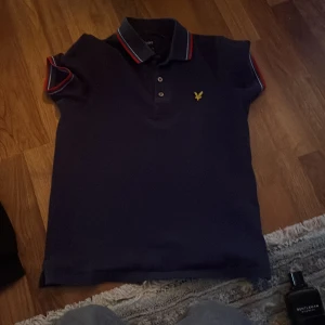 Mörkblå pikétröja från Lyle & Scott - Säljer en mörkblå pikétröja från Lyle & Scott med klassisk krage och knappar framtill. Tröjan har röda och vita ränder på krage och ärmslut samt den ikoniska gula loggan broderad på bröstet.