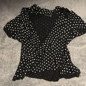 Svart prickig omlottblus - Säljer en svart omlottblus från Boohoo med vita prickar. Blusen har korta ärmar och volangdetaljer som ger en söt touch. Perfekt för en stilren look!