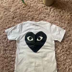 Vit t-shirt från Comme des Garçons Play - Vit t-shirt från Comme des Garçons Play med svart tryck framtill och stort svart hjärta med ögon på ryggen. Klassisk rund halsringning och korta ärmar. Perfekt för dig som gillar streetwear och stilrena plagg.