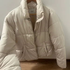 Beige pufferjacka från Jacqueline de Yong - Snygg beige pufferjacka från Jacqueline de Yong med hög krage och dragkedja framtill. Jackan har två fickor på sidorna och quiltad design som ger en modern look. Perfekt för kyligare dagar och enkel att matcha med olika outfits.