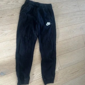 Svarta mjukisbyxor från Nike - Svarta mjukisbyxor från Nike med vit logga på vänster ben. Byxorna har resår i midjan och muddar vid bensluten. Perfekta för en avslappnad stil eller träning.