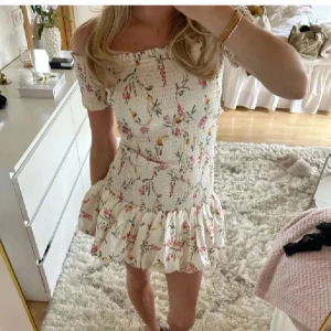 Blommig kortklänning med volang - Supersöt kortklänning med blommigt mönster och volang nertill. Klänningen har smockad överdel, fyrkantig ringning och korta ärmar. Perfekt för sommarens alla tillfällen! Använd 1-3 gånger