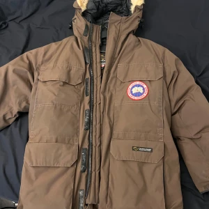 Brun dunjacka från Canada Goose med pälsdetalj - Säljer en brun dunjacka från Canada Goose med svart quiltat foder och avtagbar huva med päls. Jackan har flera stora fickor framtill, dragkedja och knappar. Perfekt för kalla dagar och riktigt varm. Kan mötas upp i Göteborg eller frakta mot betalning. Lite trasig på höger ärm i tyget, går att fixa.