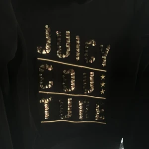 Svart velour hoodie från Juicy Couture - Svart hoodie i mjuk velour från Juicy Couture med dragkedja och huva. Framsidan har fickor och baksidan är dekorerad med guldiga paljetter som bildar texten 'Juicy Couture'. Perfekt för en avslappnad och trendig look.