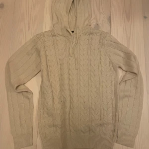 Stickad Hoodie | BRAVE SOUL. - Säljer en beige stickad hoodie från Brave Soul med kabelstickat mönster. Tröjan har huva med dragsko och långa ärmar. Perfekt för kyliga dagar och enkel att matcha med olika outfits.