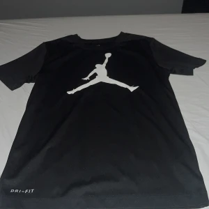 Svart Jordan Dri-Fit t-shirt - Säljer en svart t-shirt från Jordan med den klassiska Jumpman-loggan i vitt på bröstet. T-shirten är tillverkad i Dri-Fit-material som andas och passar perfekt för träning eller sport. Kortärmad och med rund halsringning. Passar 150-160 cm lång