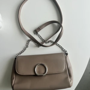 Beige axelväska med kedjedetalj - Säljer en stilren beige axelväska med silverfärgad kedja och rund metallring på locket. Perfekt storlek för det viktigaste och passar till många olika outfits.