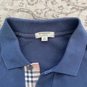 Blå pikétröja från Burberry - Säljer en klassisk blå pikétröja från Burberry i slim fit-modell. Tröjan har korta ärmar, krage och knäppning med Burberrys ikoniska rutiga detalj. Tillverkad i bomull och har en liten broderad logga på bröstet. Har en mindre fläck som man knappt ser under högra bröstet, se sista bilden.