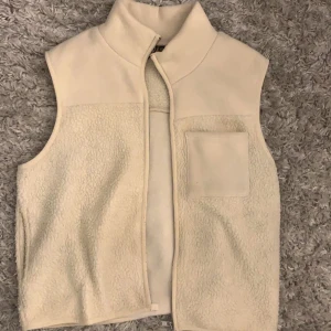 Beige fleeceväst med hög krage - Mysig beige fleeceväst med hög krage och en bröstficka. Perfekt att slänga på sig över en hoodie eller tröja när det är kyligt ute. Enkel och stilren design med en enkel dragkedja framtill