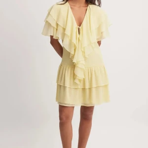 Gul volangklänning  - Söt gul klänning med volanger och knyt framtill. Djup v-ringning och dubbla volanglager upptill samt volangkjol. Perfekt för somriga dagar och midsommarfirande. Från nakd, använd en gång❤️strl 36 men passar 36/38