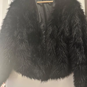 Svart fuskpälsjacka - Säljer en svart, fluffig fuskpälsjacka med croppad passform. Jackan har lång ärm och är helsvart. Perfekt för dig som vill sticka ut och hålla dig varm under kyliga dagar.