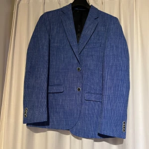 Snygg blå kavaj - Snygg blå kavaj från ”Luxury Brand” med diskret strukturmönster. Kavajen har två knappar framtill, fyra knappar vid ärmslut och är helfodrad med ljusblått foder. Perfekt för bal eller fest. Storlek 102 vilket är 52 men något längre armar.