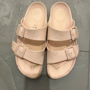 Beige sandaler med spännen - Säljer ett par beige sandaler med två breda remmar och stora spännen. Skorna har en platt sula och är tillverkade i syntetmaterial. Perfekta för varma dagar och enkel att ta på sig.