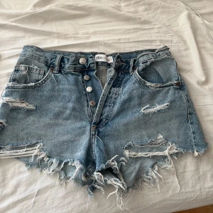 Blå jeansshorts med fransar från Zara - Säljer ett par blå jeansshorts från Zara med  slitna detaljer och fransig kant. Shortsen har knappar framtill och klassiska fickor både fram och bak. Perfekta för sommaren!