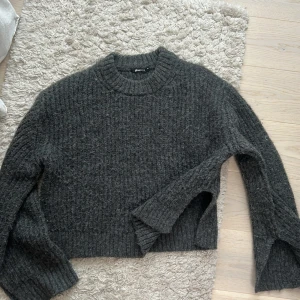 Mörkgrå stickad tröja från Gina Tricot - Mysig mörkgrå stickad tröja från Gina Tricot med grov ribbstickning och rund halsringning. Tröjan har långa ärmar och en något croppad passform. Har slits på sidan av armarna och midjan.