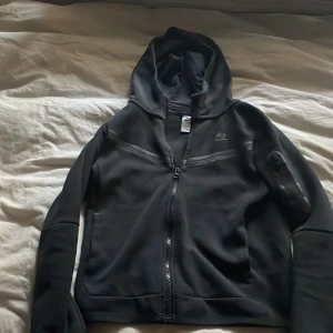 Svart hoodiejacka från Nike ( Nike teech) - Säljer en svart hoodiejacka från Nike med dragkedja framtill och huva. Jackan har långa ärmar och en sportig look, perfekt för träning eller chill. Klassisk Nike-logga på bröstet och praktiska fickor.