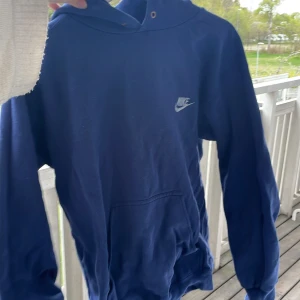 Blå hoodie från Nike - Mörkblå hoodie från Nike med klassisk logga på bröstet. Tröjan har huva och magficka, samt ribbade muddar vid ärmslut och nederkant. Perfekt för en avslappnad och sportig stil.