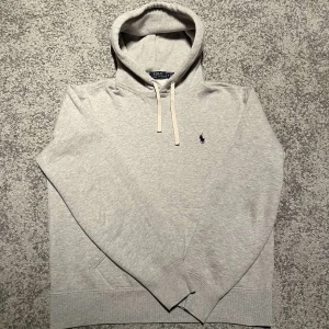 Ralph Lauren hoodie  - Säljer en stilren grå hoodie från Polo Ralph Lauren med klassisk logga på bröstet. Tröjan har en justerbar huva med vita snören och en praktisk magficka. Perfekt för en avslappnad look.