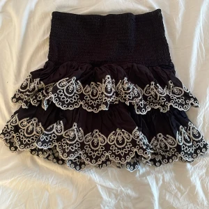 Mini kjol - Supersöt svart kortkjol med bred smock i midjan och två lager volanger. Kjolen har vita broderade spetsdetaljer längs volangkanterna som ger en unik och festlig look. Perfekt att styla med en enkel topp för en trendig outfit. Man kan göra midjebandet kortare om man vill. Midjebandet är även stretchigt. Kjolen passar mellan XS till M. Men skulle passformen är menad för XS-S. Skriv via funderingar och mer bilder💕