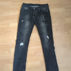 Dsquard 2 Jeans - Säljer ett par svarta jeansbyxor med slitningar och färgstänk för en cool, använd look. Byxorna har klassisk femficksdesign, knapp- och dragkedjegylf samt raka ben. Perfekta för dig som gillar streetstil.