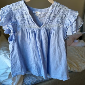 Ljusblå blus från gina - Supersöt ljusblå blus med volangärmar och v-ringning. Blusen har fina rynkade detaljer upptill och är tillverkad i ett lätt och luftigt tyg, perfekt för varmare dagar.