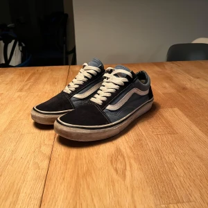 Blå och svarta Vans Old Skool sneakers - Säljer ett par klassiska Vans Old Skool sneakers i blått och svart med vit sula och vit sidostripes. Skorna har snörning och den ikoniska gummisulan med rutmönster. Perfekta för en avslappnad och stilren look. Sulan inne i skon är lite sönder men går att byta