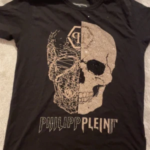 Svart t-shirt med döskalle från Philipp Plein - Cool svart t-shirt från Philipp Plein med en stor döskalle framtill, där ena halvan är glittrig och andra halvan har ett grafiskt mönster. T-shirten har rund hals och korta ärmar. Märkeslogga på ryggen.