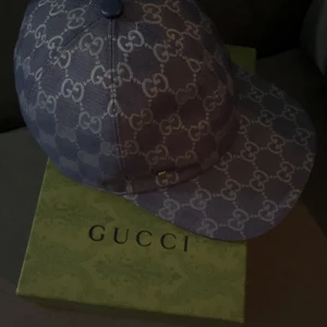 Grå keps från Gucci med monogram - Säljer en snygg grå keps från Gucci med klassiskt GG-monogram i vitt. Kepsen har böjd skärm och justerbar passform. Perfekt för dig som gillar exklusiva accessoarer och vill sticka ut med stil.