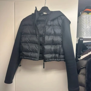 Svart croppad dunväst med huva - Säljer en svart croppad dunväst med huva och långa ärmar. Västen har dragkedja framtill och är i en sportig, modern stil. Perfekt för lager-på-lager och streetwear. Snygg och enkel att matcha med olika outfits.