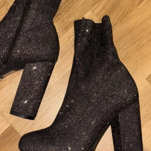Svarta glittriga klack skor  - Säljer ett par svarta stövletter med glittrigt material och hög klack. Skorna har en rejäl platåsula och dragkedja på sidan. Perfekta för att sticka ut och ge extra höjd till din outfit.