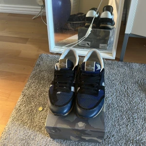 Valentino Garavani sneakers i svart och blå - Säljer ett par snygga Valentino Garavani sneakers i svart och blå med vita detaljer. Skorna har svart snörning, rund tå och platt sula. Ovandelen är i en mix av skinn och textil, med märkets logga på plösen. Perfekta för dig som vill ha något exklusivt och stilrent. Box kommer med, även påse, snören. skorna är nästan aldrig använda, så väldigt bra skick!