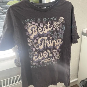 Svart t-shirt med tryck från Lindex - Svart t-shirt från Lindex med stort färgglatt tryck framtill med texten 'Best Thing Ever', blommor och fjärilar. T-shirten har rund halsringning och korta ärmar. Perfekt för dig som gillar statement-plagg med coolt motiv.