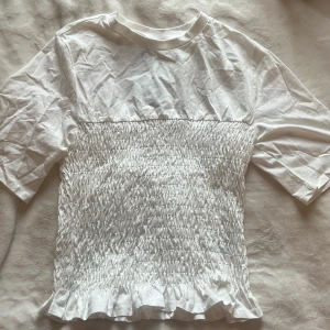 Vit smockad topp med volang från H&M Divided - Säljer en vit topp från H&M Divided med smockad midja och volangkant nedtill. Toppen har korta ärmar och är tillverkad i bomull med 25% återvunnet material. Perfekt för en fräsch och söt look!